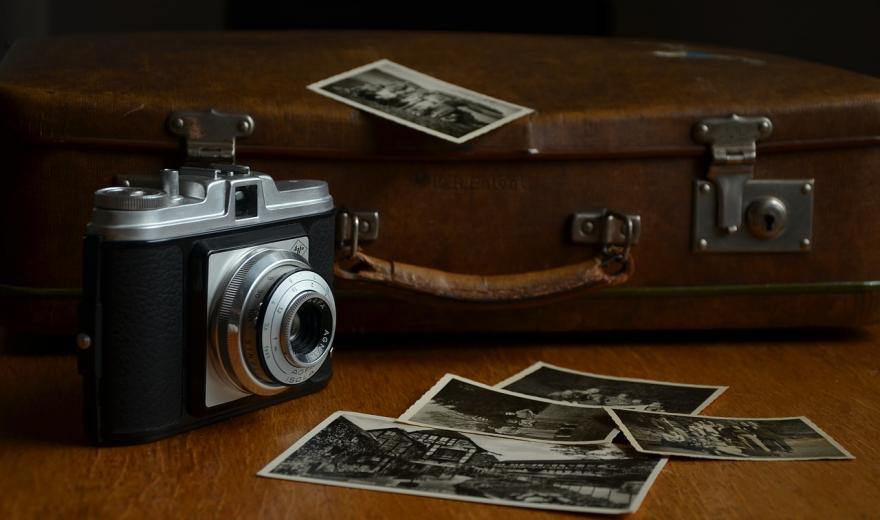 Een bruin tafelblad waarop in de achtergrond een oude bruine koffer ligt met één zwartwit foto erop, linksvoor een analoge Agfa Isola camera staat met rechts daarnaast vier over elkaar liggende zwartwit foto's.