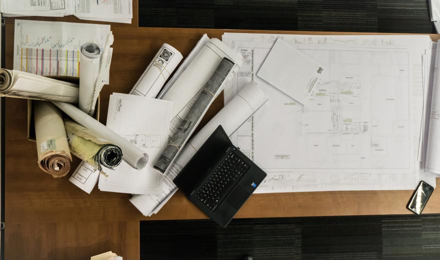 Bovenaanzicht van een notenhouten bureautafel bedekt met opgerolde bouwtekeningen in een doos, diverse opgerolde en opengerolde bouwtekeningen, een opengeslagen laptop en een mobiele telefoon.