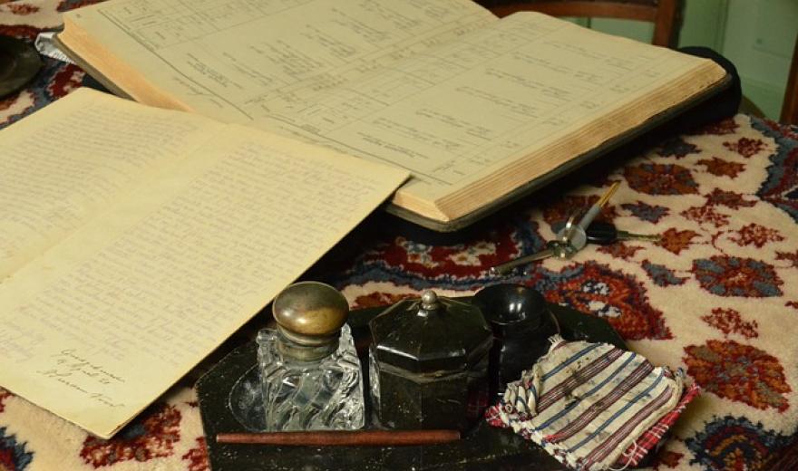 Op een tafelkleed met bonte patronen staat een tableau met inktpot, een doekje en een kroontjespen voor een opengeslagen registerboek  met losliggend vel papier.